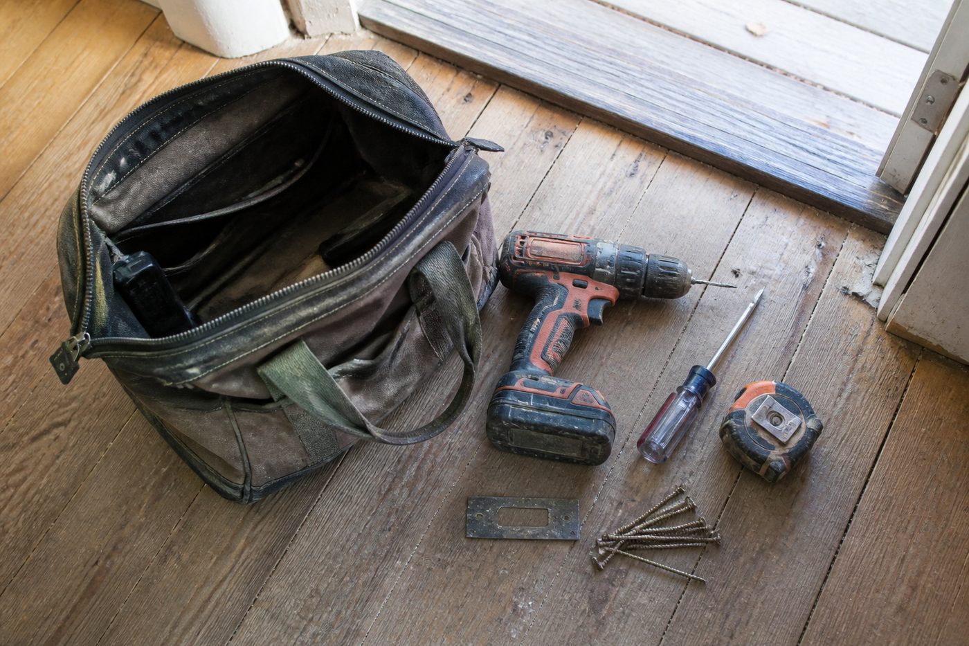 An open tool bag beside a front door with a drill, a strike plate and long screws laid out